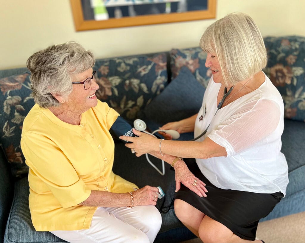Older woman receiving a blood pressure check and speaking with a care professional in a comfortable living room.