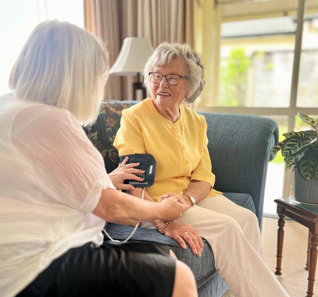 Older woman in a yellow blouse receiving a blood pressure assessment from a smiling care professional.